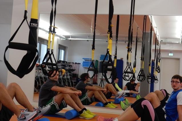 Mehrere Personen sitzen in einem Fitnessraum auf Rollen, w&auml;hrend TRX-B&auml;nder von der Decke h&auml;ngen.