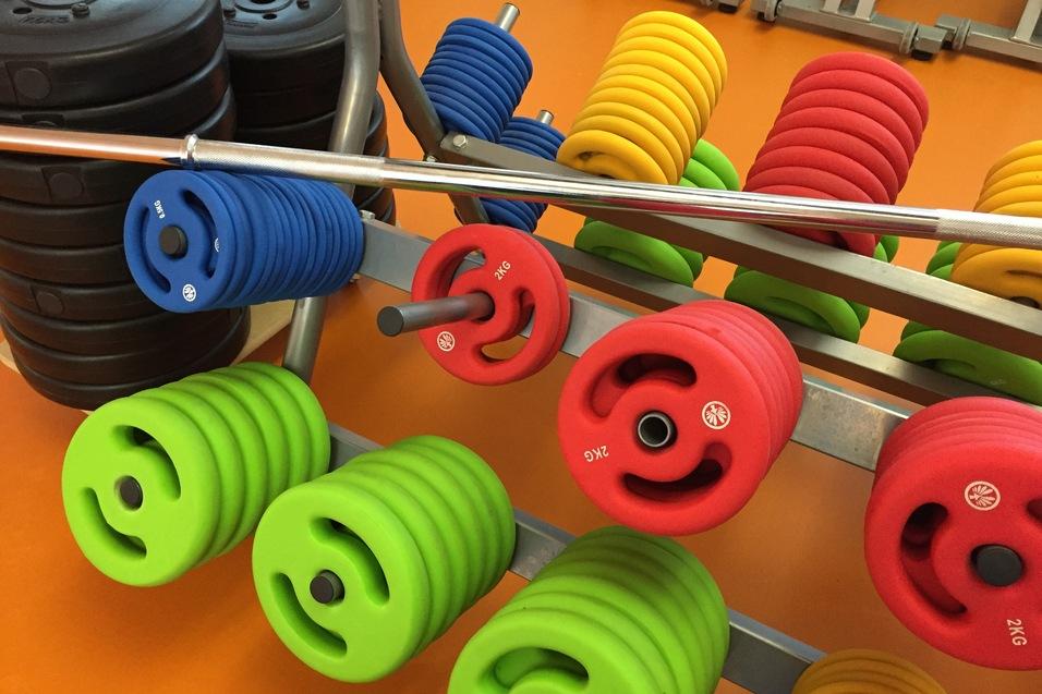Reihe bunter Fitnessgewichte in verschiedenen Größen, zusammen mit einer Langhantel auf einem Fitnessstudio-Boden.