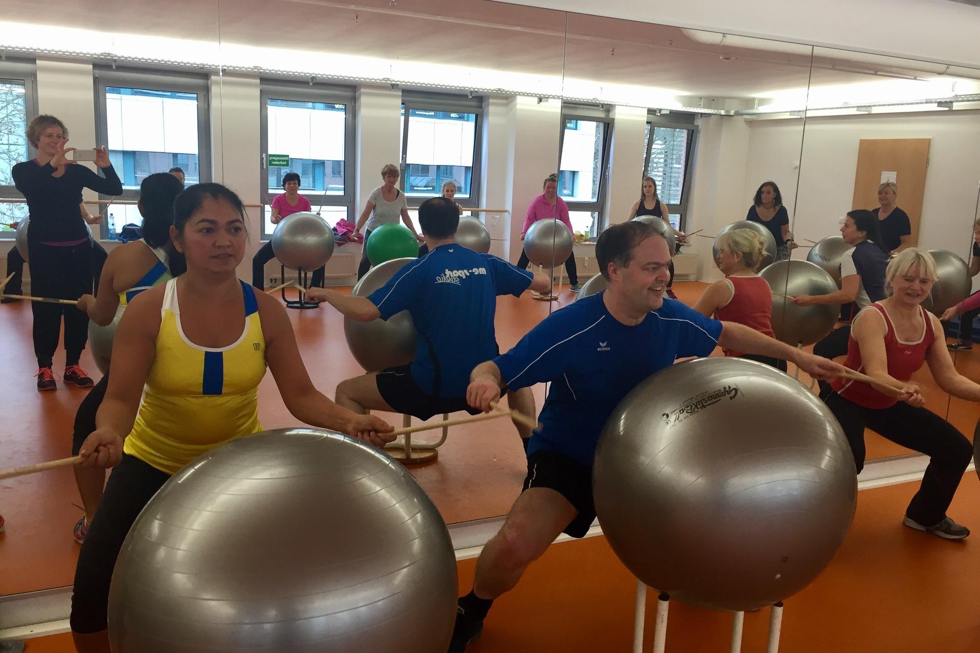 Gruppe von Personen in einem Fitnessraum, die mit Gymnastikbällen und Sticks an einer Übung teilnehmen.