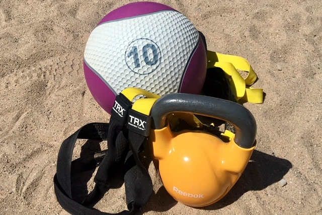 Fitnessgerät auf Sandboden: Ein lilafarbener Medizinball, ein TRX-Gurt und eine orangefarbene Kettlebell.