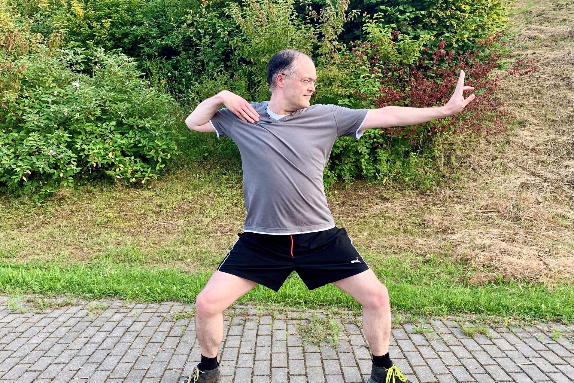 Mann in Sportkleidung steht in einer dynamischen Pose auf einem gepflasterten Weg, umgeben von Grünflächen.