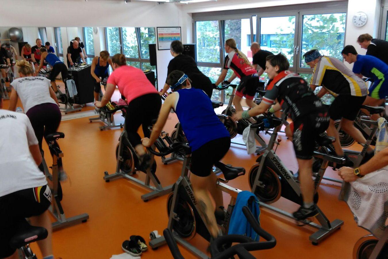 Gruppe von Menschen beim Indoor-Cycling in einem Fitnessstudio mit mehreren Spinning-Bikes und großen Fenstern.