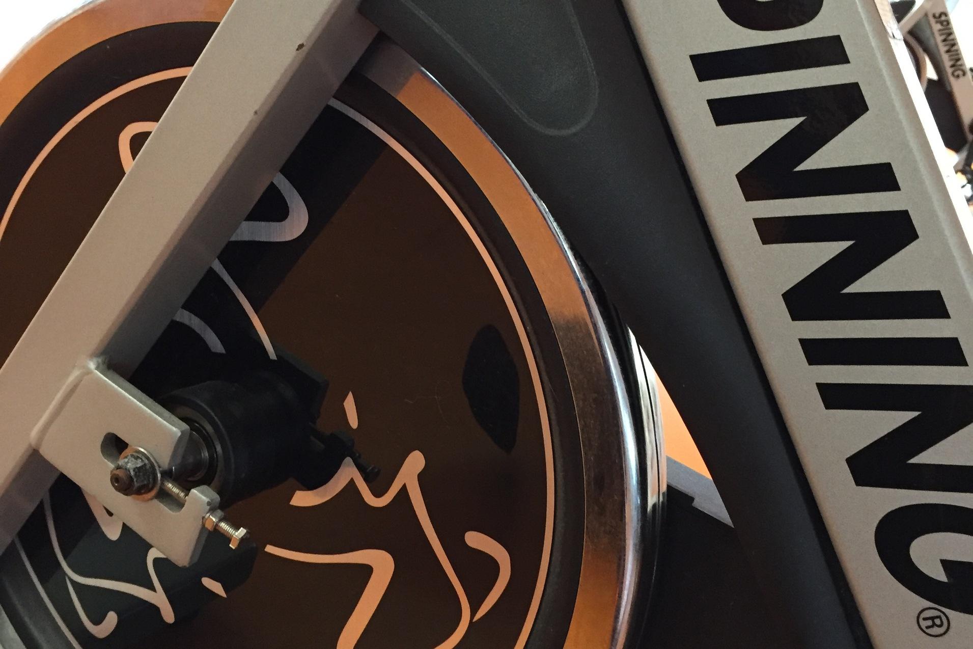 Detailansicht eines Spinning-Bikes mit dem Logo auf dem Schwungrad, vor einem orangefarbenen Untergrund.