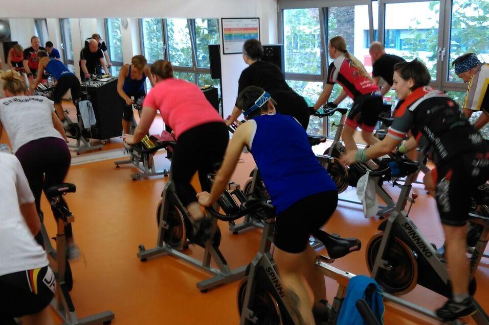 Gruppe von Menschen beim Indoor-Cycling in einem Fitnessstudio mit mehreren Spinning-Bikes und großen Fenstern.