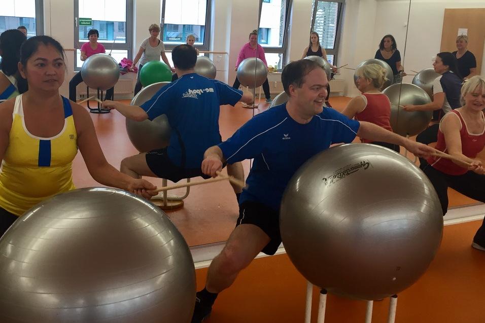 Gruppe von Personen in einem Fitnessraum, die mit Gymnastikbällen und Sticks an einer Übung teilnehmen.