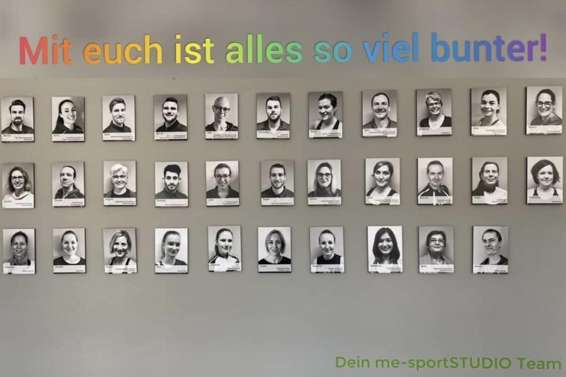 Wand mit Schwarz-Wei&szlig;-Portr&auml;ts von Teammitgliedern, dar&uuml;ber der Text &bdquo;Mit euch ist alles so viel bunter!&ldquo; in bunten Buchstaben.