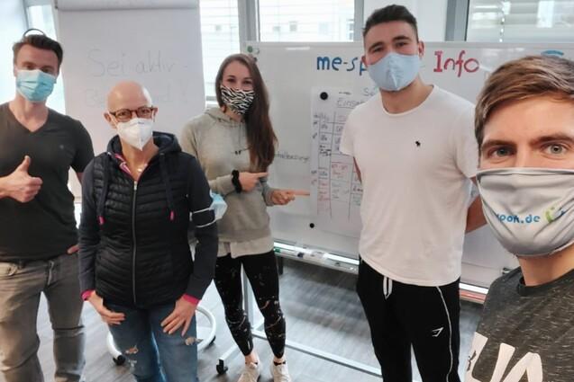 Gruppenfoto von f&uuml;nf Personen in Masken, die vor einem Flipchart in einem modernen Raum stehen.