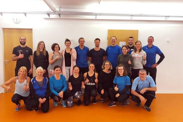 Gruppe von 20 Personen in Sportkleidung, die im Raum stehen und lächeln, einige zeigen Daumen hoch.