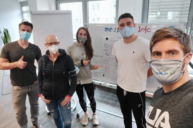 Gruppenfoto von fünf Personen in Masken, die vor einem Flipchart in einem modernen Raum stehen.