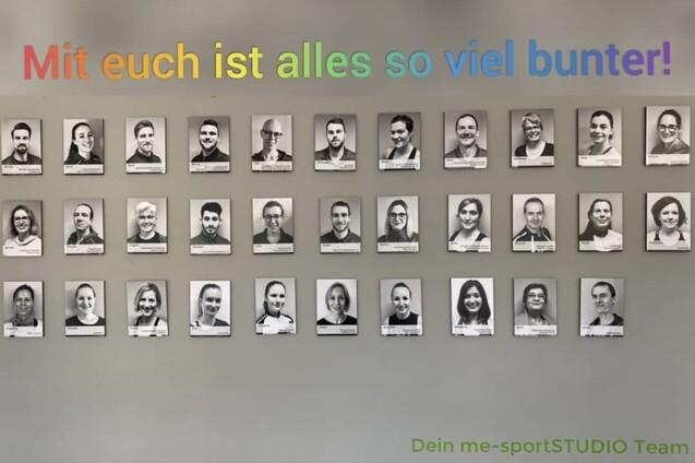 Wand mit Schwarz-Weiß-Porträts von Teammitgliedern, darüber der Text „Mit euch ist alles so viel bunter!“ in bunten Buchstaben.