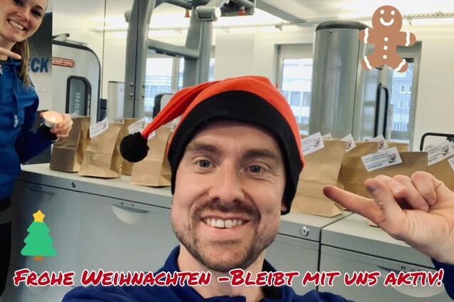 Selbstporträt eines Mannes mit Weihnachtshut und einer Frau im Hintergrund, vor Tüten in einem Fitnessstudio.