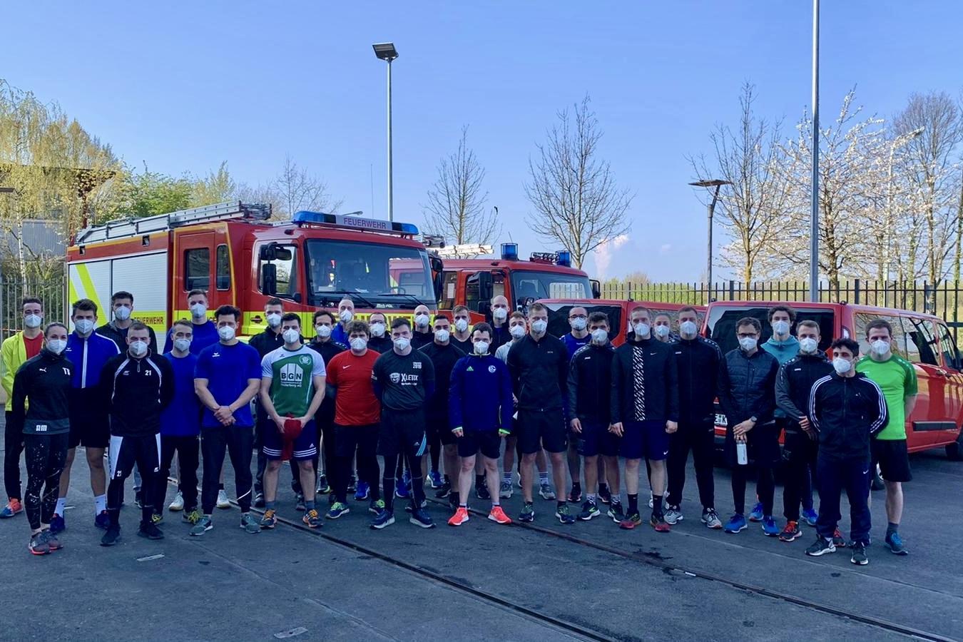 Gruppe von 24 Personen in Sportkleidung und Masken, vor zwei Feuerwehrfahrzeugen und blühenden Bäumen auf einem Platz.