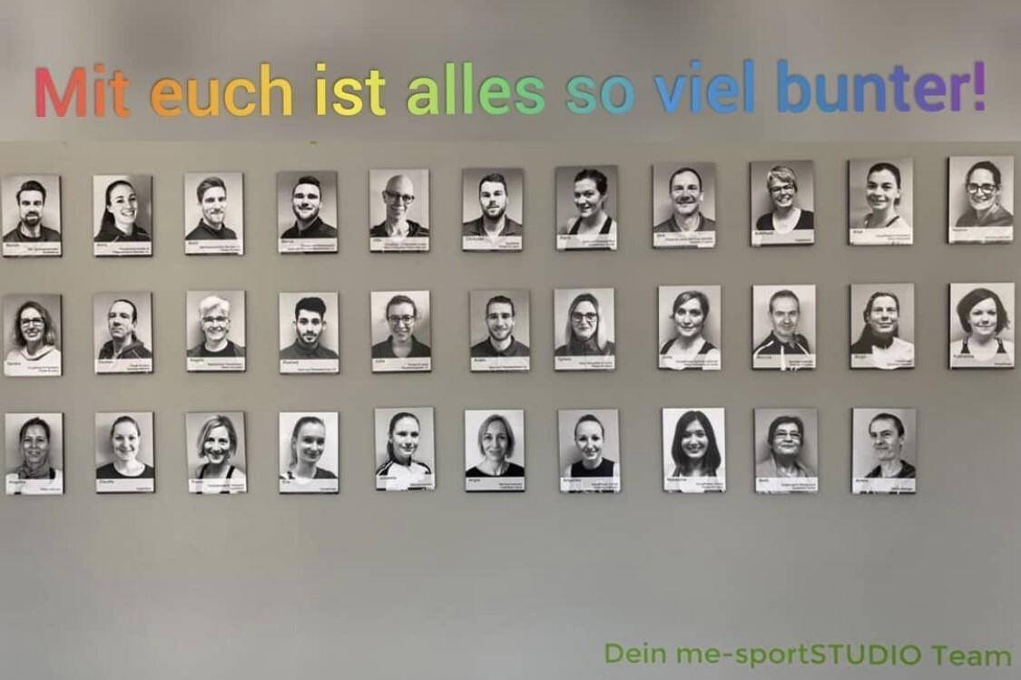 Wand mit Schwarz-Weiß-Porträts von Teammitgliedern, darüber der Text „Mit euch ist alles so viel bunter!“ in bunten Buchstaben.