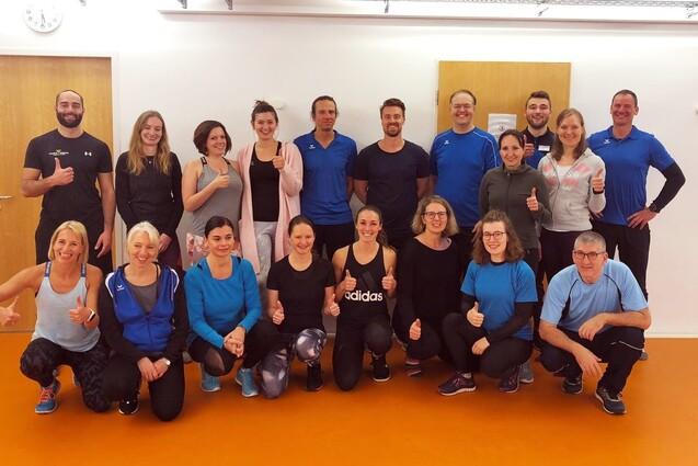 Gruppe von 20 Personen in Sportkleidung, die im Raum stehen und l&auml;cheln, einige zeigen Daumen hoch.