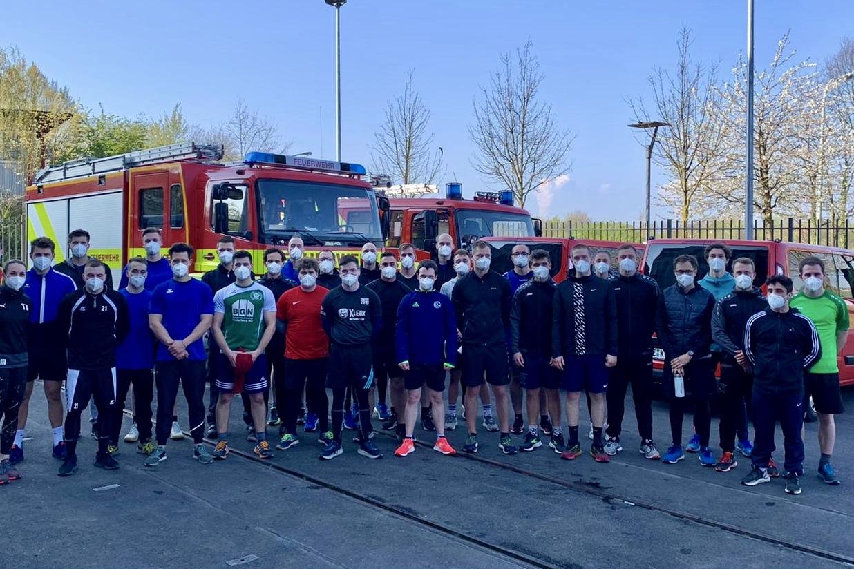 Gruppe von 24 Personen in Sportkleidung und Masken, vor zwei Feuerwehrfahrzeugen und bl&uuml;henden B&auml;umen auf einem Platz.